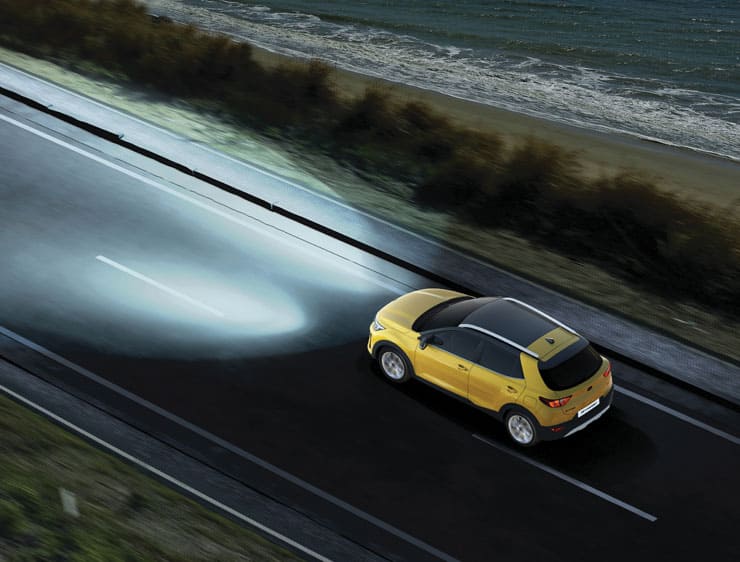 A yellow color Kia Stonic SUVwith lights on in a beachside road